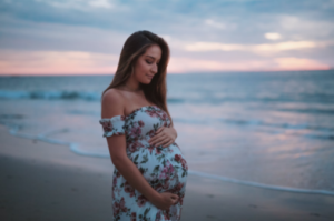 Pregnant woman in floral dress standing again the sunset on the beach