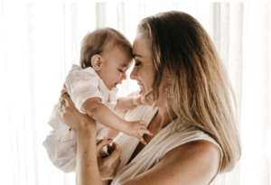 A blonde woman in a beige dress holding a newborn in a white outfit