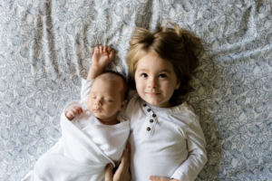 a blonde toddler and bald newborn on top of floral sheets