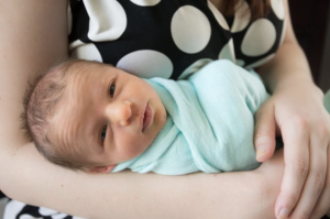 female in polka dot dress holding a newborn in a pal green blanket