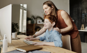 Red haired woman in a rust colored outfit is helping a red haired boy in a blue shirt at the computer