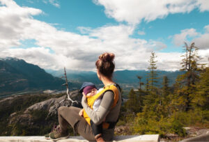 Woman holding baby in a yellow carrier overlooking mountains and trees. 